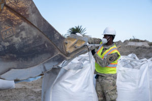 Louisiana National Guardsman assigned to the 225th Engineer Brigade hooks straps of a 3-by-3 foot sack to an excavator bucket filled with up to 4,000 pounds of sand to emplace in one of twelve levee breaches, Grand Isle, Sept 24, 2021. To date, the 225th has emplaced more than 1,100 sacks of sand along the coastline levees in Grand Isle. (U.S. Army National Guard photo by Staff Sgt. Josiah Pugh)